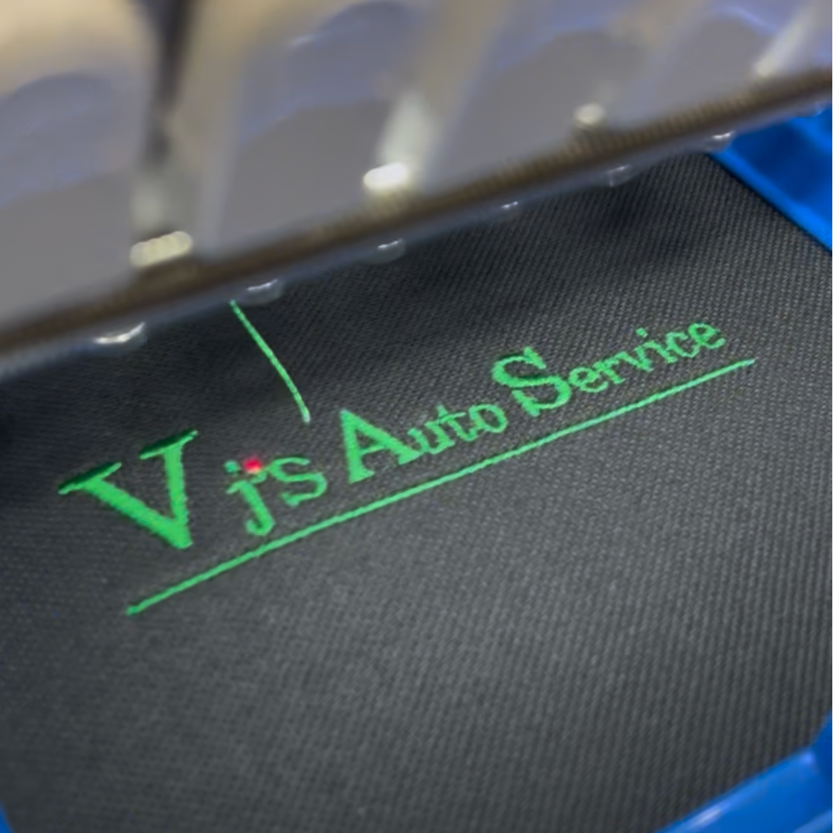 A close-up of a dark gray fabric surface with green embroidered text reading 'Vjs Auto Service' under a green underline, with part of a blue and white machine visible around it.
