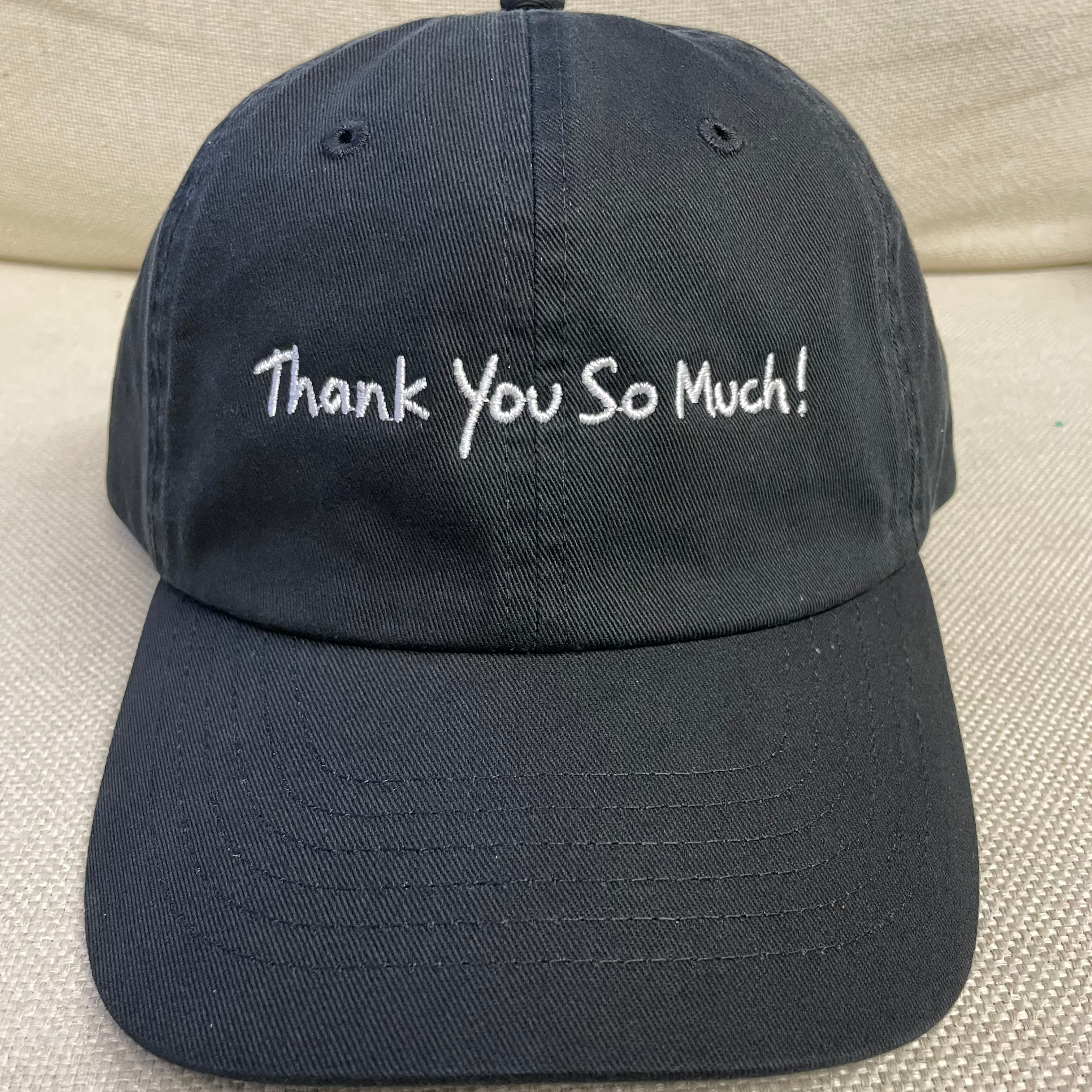A dark navy blue baseball cap with 'Thank You So Much!' embroidered in white script across the front, resting on a textured beige fabric surface.