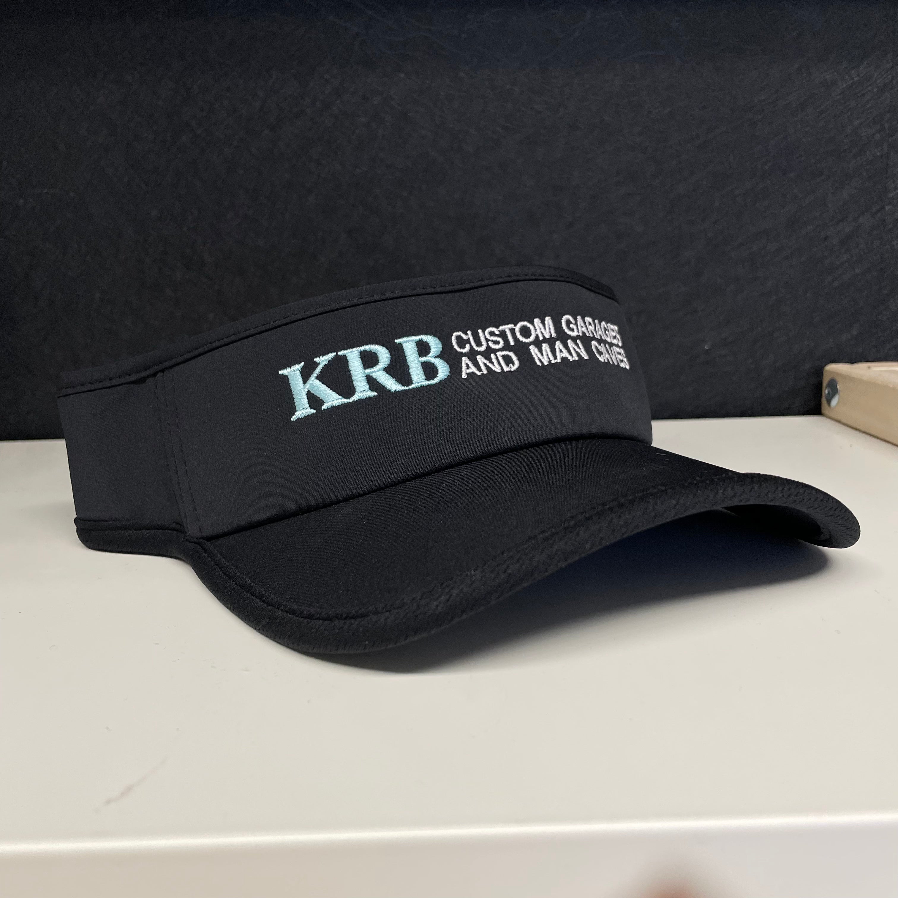 A black visor-style cap with embroidered text reading 'KRB Custom Garage and Man Caves' in light blue and white, resting on a white surface against a dark textured background.