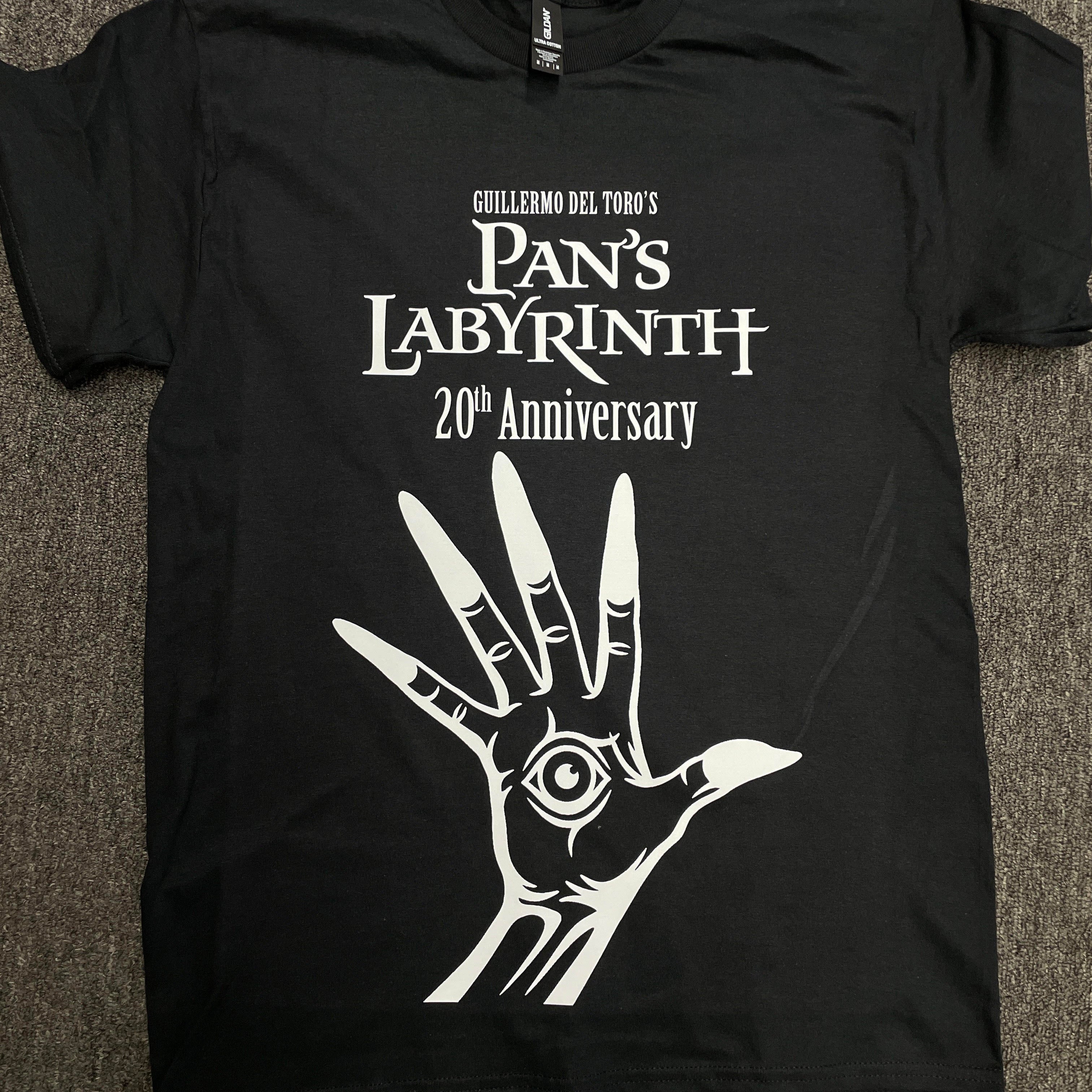 A black short-sleeve graphic T-shirt laid flat on a gray carpeted surface. The shirt features white text reading 'CHILEBIM DEL TONO'S Pan's Labyrinth 20th Anniversary' above a large white hand graphic with an eye in the center.