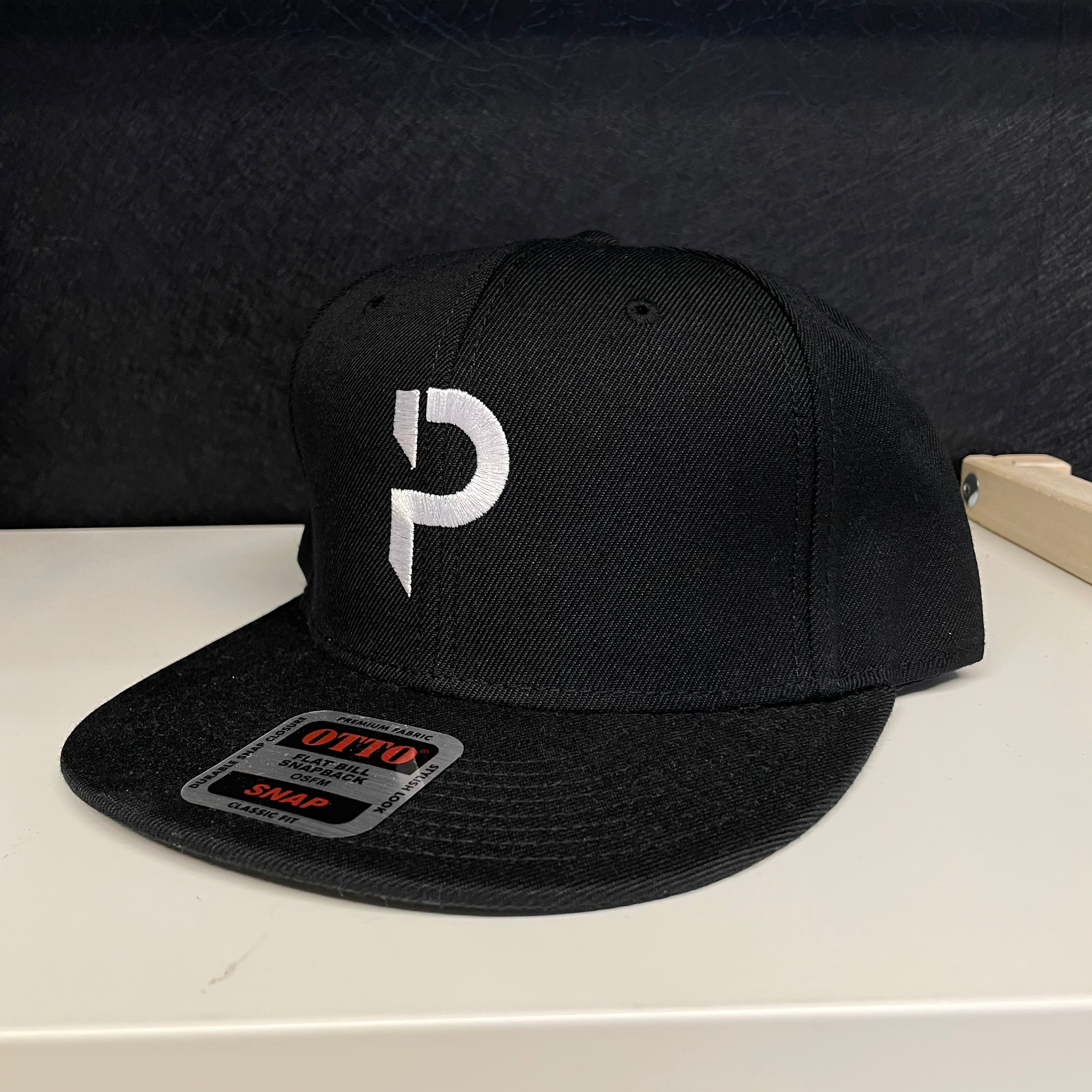 A black flat-brim snapback cap with a white stylized 'P' logo embroidered on the front, resting on a white surface against a dark textured background.